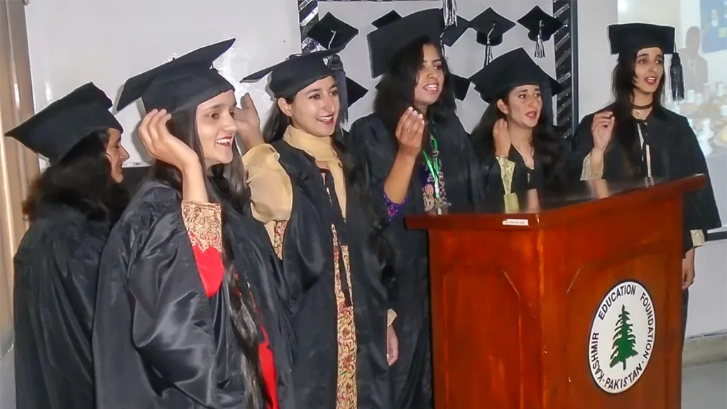 Young women in black graduation gowns and caps beside a podium with the KEF emblem