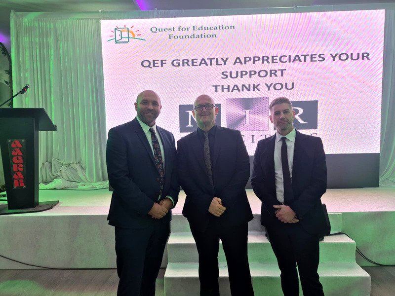 Three men in suits standing in front of a screen reading QEF greatly appreciates your support, thank you