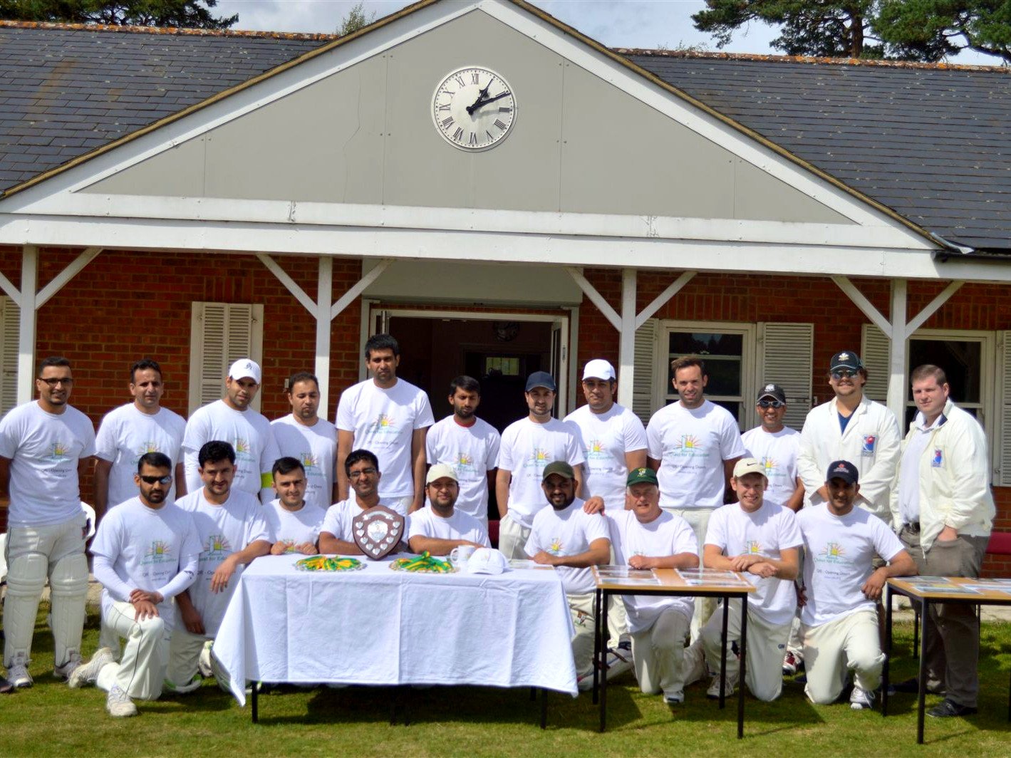 Cricket4Education, June 2014 Cricket teams in Quest for Education t-shirts posing in front of a pavilion with a shield trophy and medals on a table.