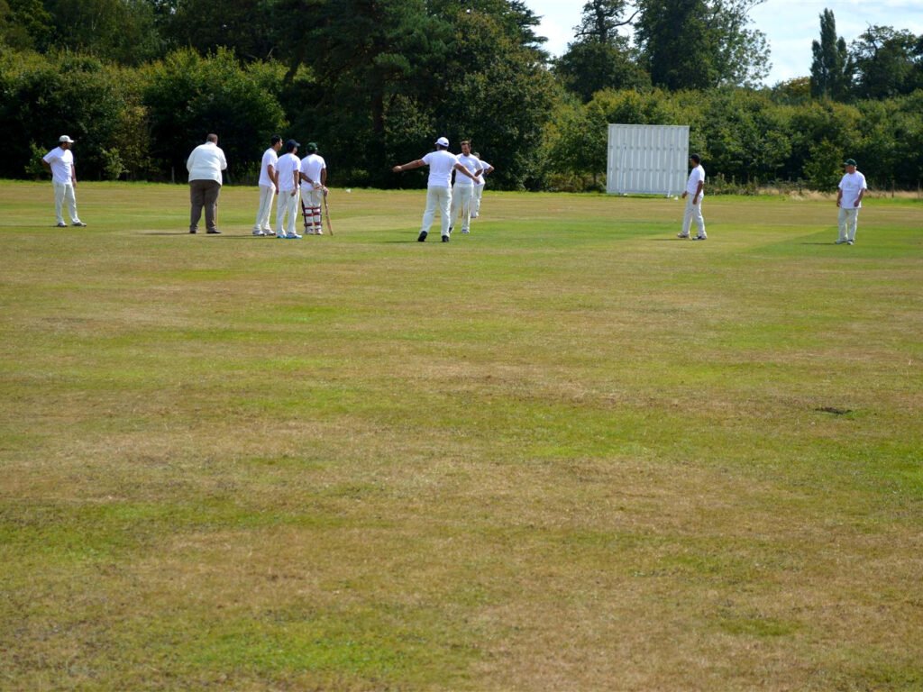 Cricketers in white on a large grass field with a sightscreen and trees behind