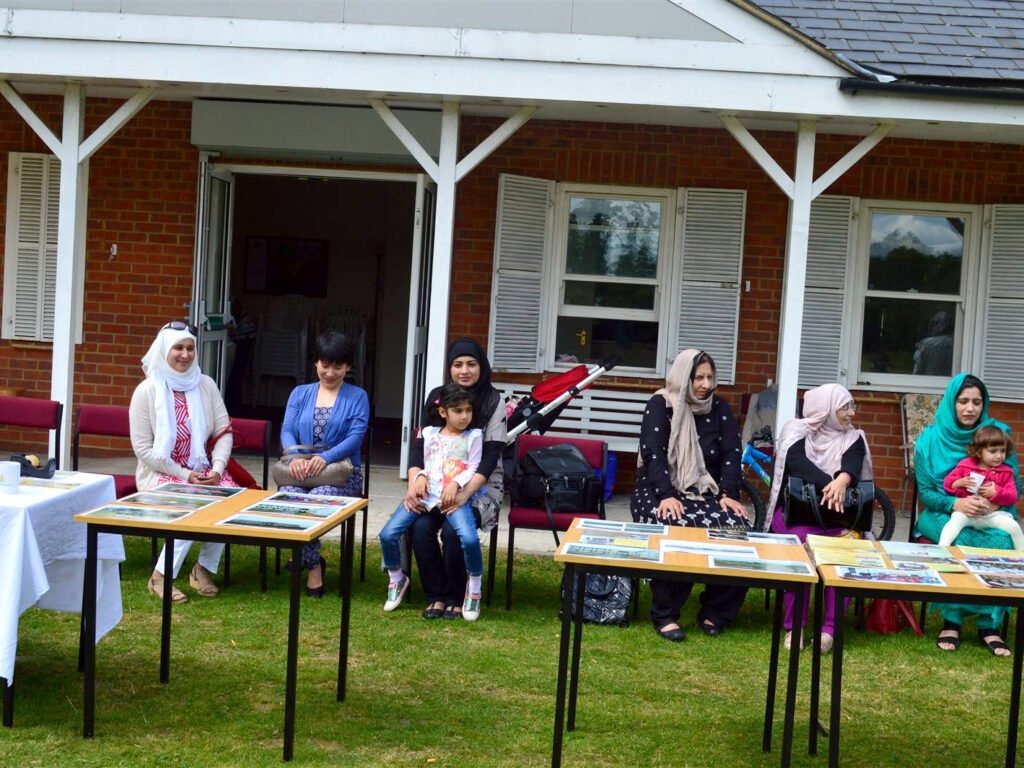 Women and children seated behind tables of displayed photographs at a cricket pavilion