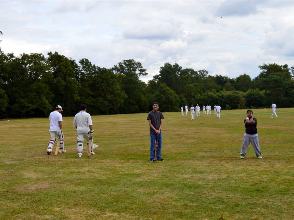 Two boys holding cricket bats on the outfield while batsmen walk past