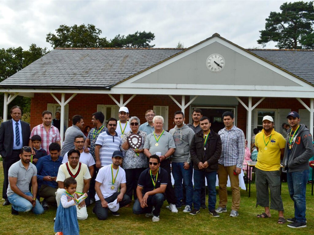 Large group of players and supporters with medals and a shield in front of a pavilion