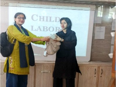 Two young women in front of a projected slide titled Child Labour, one handing the other a bag