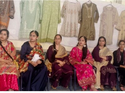 A row of women in colourful regional dress seated in front of a display of embroidered clothes