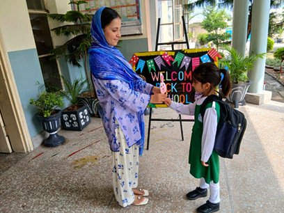 A teacher greeting a young girl with a school bag beside a Welcome back to school sign