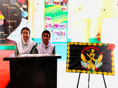 Two girls in white headscarves seated at a desk beside a decorated board