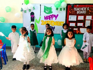 Young children in white and green outfits in front of a Happy Independence Day display