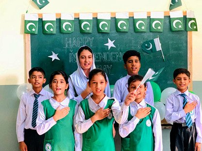 Pupils in green and white uniform with hands on their hearts in front of a chalkboard decorated with flags