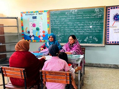 A teacher talking with a parent and child at a classroom desk beside a chalkboard