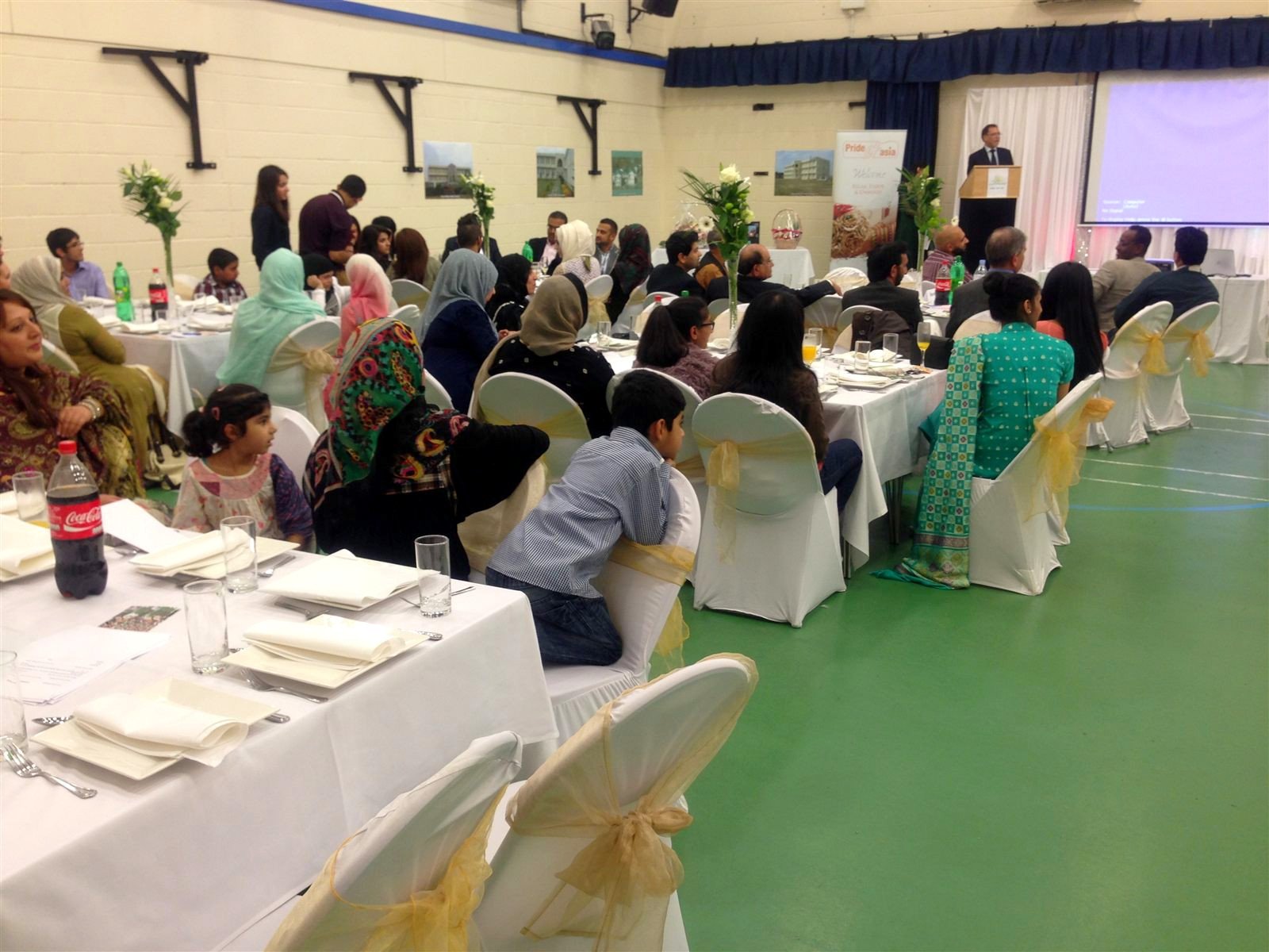 Families with children at dinner tables in a hall, speaker at the far end.