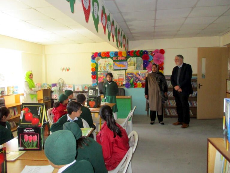 A pupil presents to her class in a school library while visitors and staff look on