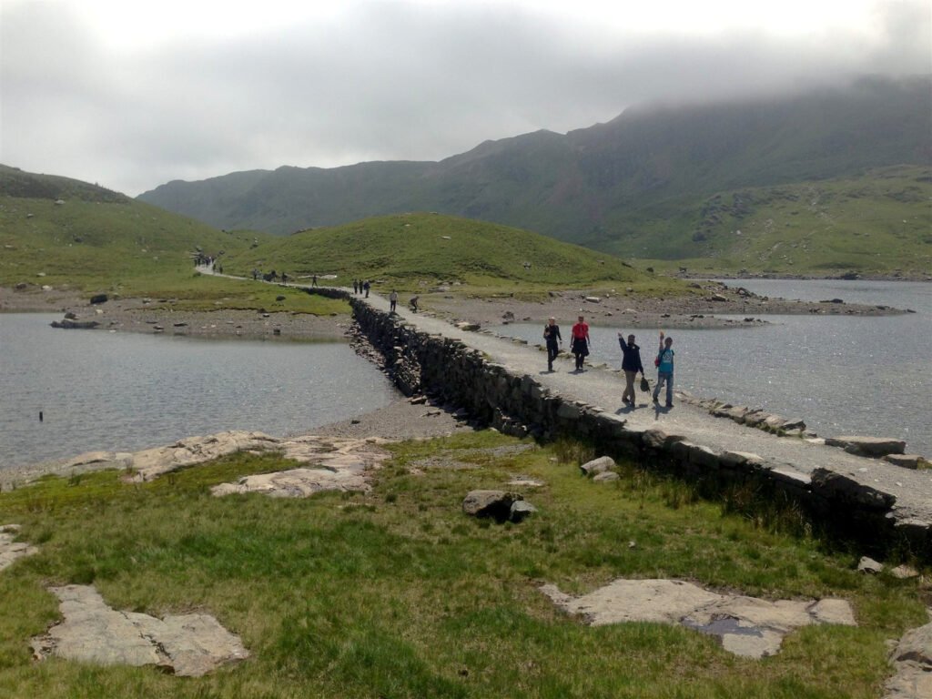 Walkers waving as they cross a stone causeway over a mountain lake